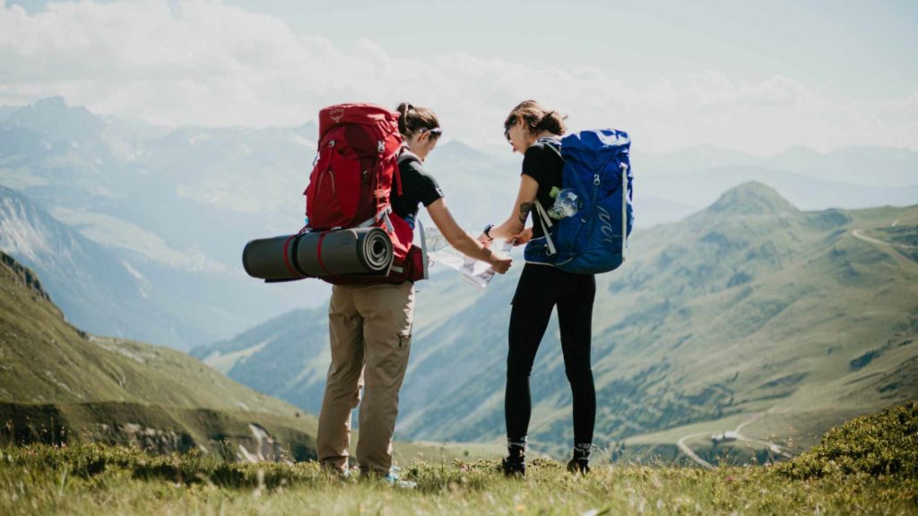 Deux randonneurs avec de grands sacs à dos consultent une carte au sommet d’une montagne, entourés de paysages verdoyants et de vallées sous un ciel dégagé.