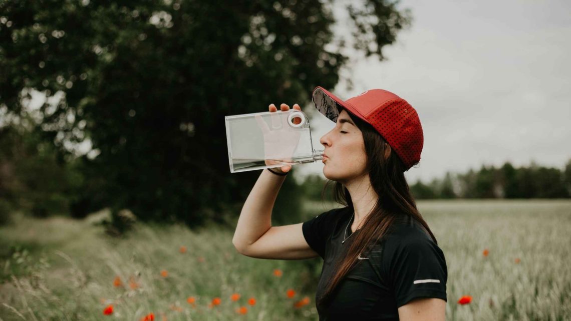 Personne portant une casquette rouge buvant de l’eau dans une gourde plate, debout dans un champ fleuri en été.