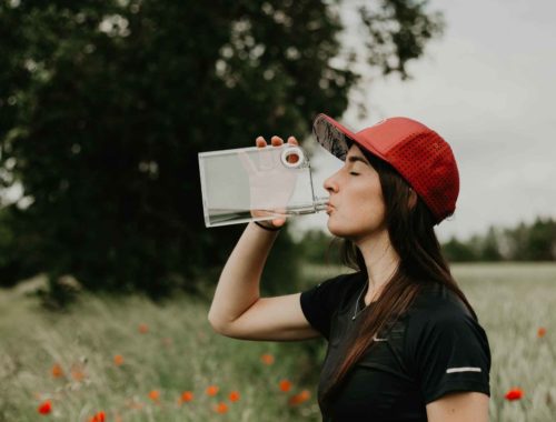 Personne portant une casquette rouge buvant de l’eau dans une gourde plate, debout dans un champ fleuri en été.