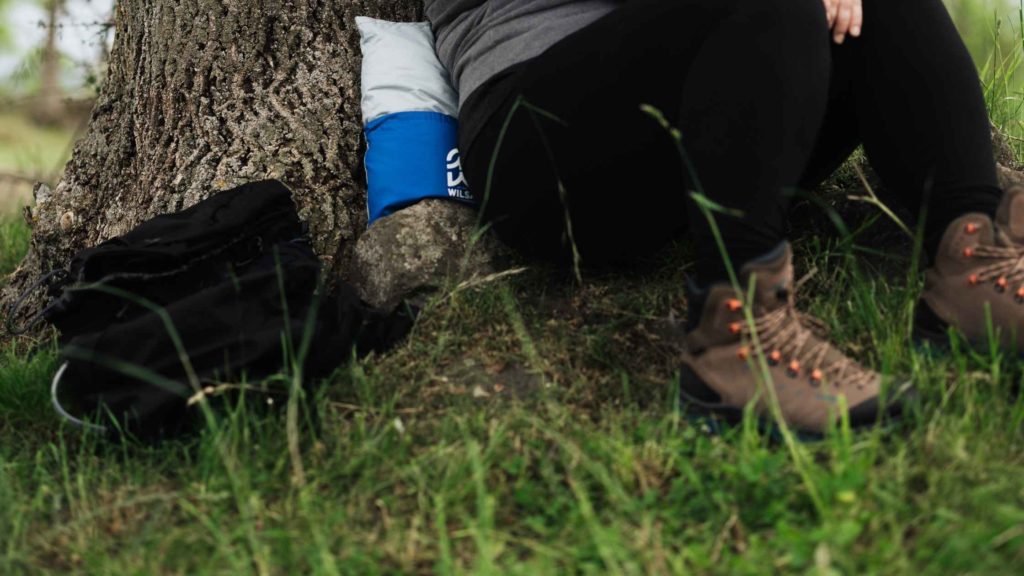 Personne assise au pied d’un arbre, en tenue de randonnée, appuyée sur un coussin bleu et blanc, avec des chaussures de marche et un sac à dos noir posé à côté.